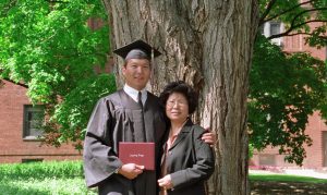 Lewis in a cap and gown holding his diploma poses for a photo under a tree with his mom.